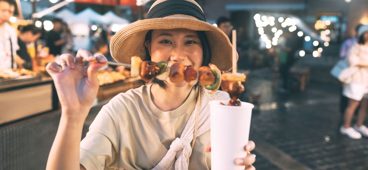 Happy young asian foodie woman eating bbq grilled skewers at outdoor night market street food vendor