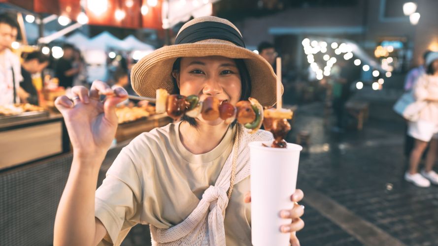 Happy young asian foodie woman eating bbq grilled skewers at outdoor night market street food vendor